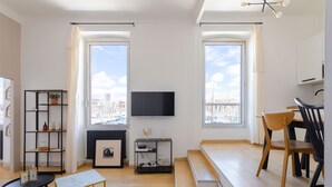 Ett modernt vardagsrum med en TV, trägolv, ett svart och guldinramat soffbord, en svart bokhylla och en matplats med trästolar.