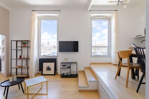 A modern living room with a TV, wooden flooring, a black and gold coffee table, a black bookshelf, and a dining area with wooden chairs.