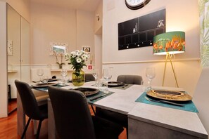 Un'area pranzo con un tavolo apparecchiato per quattro persone, sedie, un vaso con fiori, una sveglia al muro e una lampada.