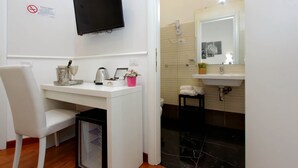 A modern bathroom with a white sink, mirror, and toilet. There's a TV mounted on the wall and a small table with a kettle and flowers.