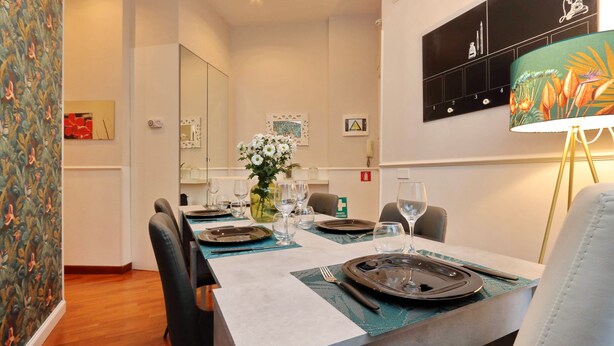 A dining area with a table set for four, including plates, cutlery, and glasses. There is a vase with flowers on the table, and the room has a modern design with a clock on the wall and a decorative lamp.