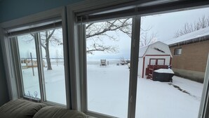 Uitzicht door een raam op een besneeuwde omgeving met een houten terras, een tuinhuis en een bank.