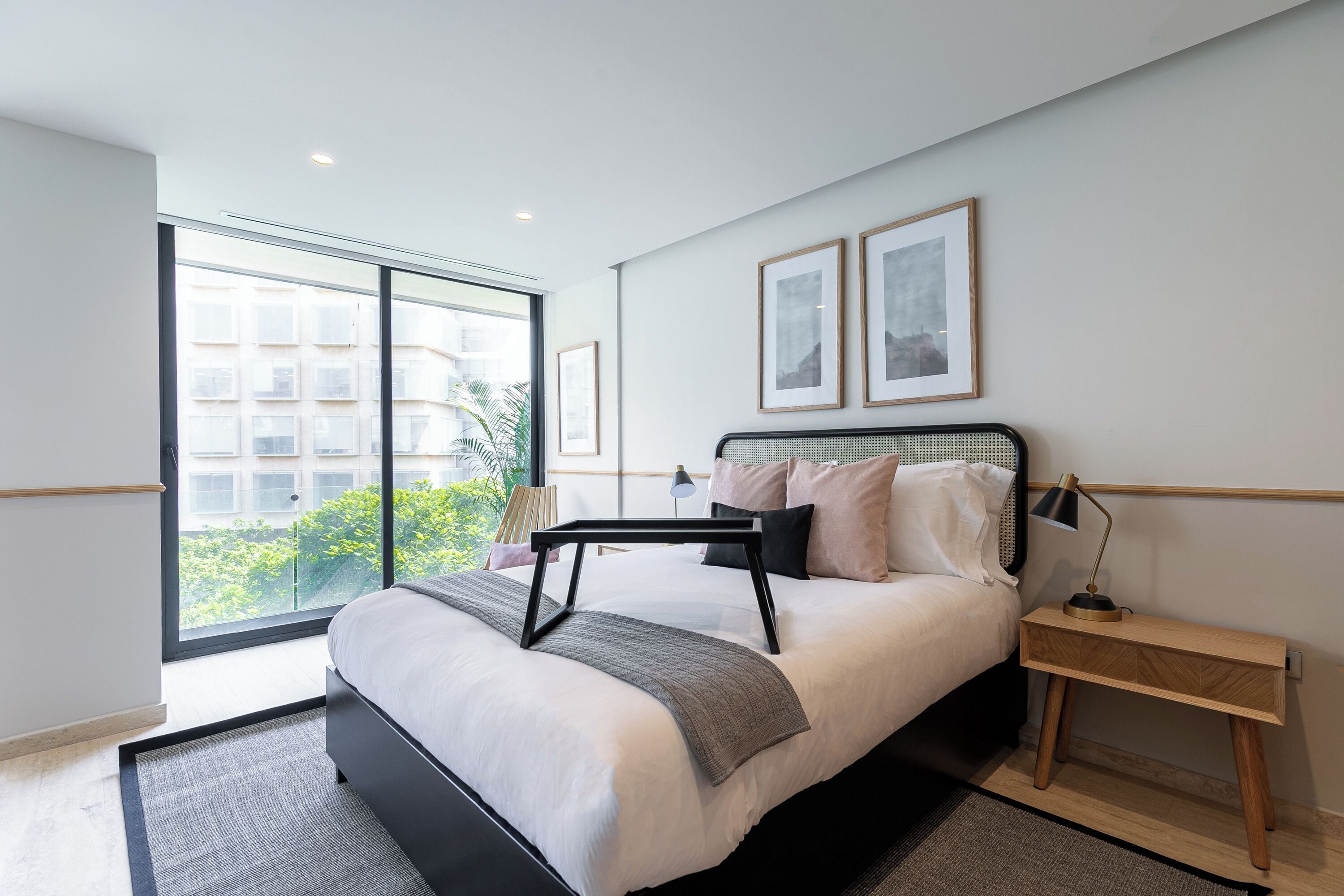 A modern bedroom with a large window, a bed with a black frame, a bedside table with a lamp, and framed pictures on the wall.