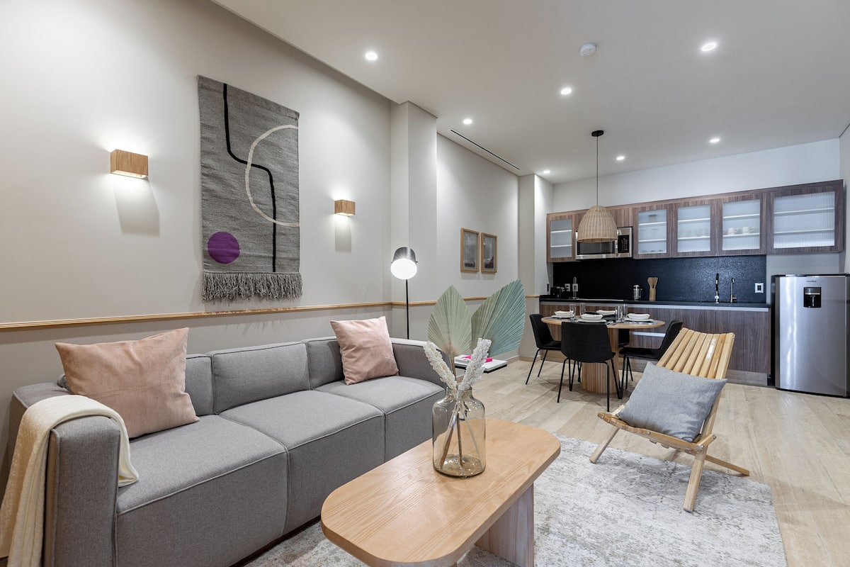 Una sala moderna con un sofá gris, una mesa de centro de madera y una cocina al fondo.
