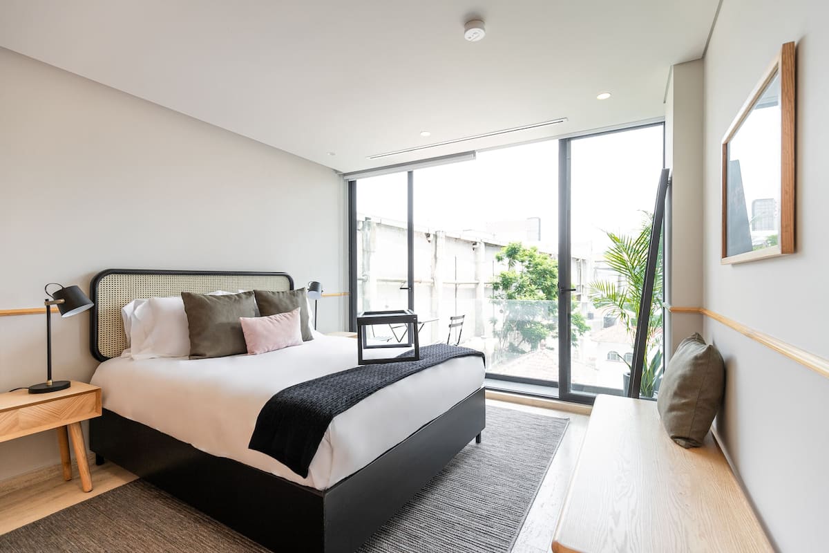 Un dormitorio moderno con una cama grande, mesitas de noche, un escritorio y vistas al exterior.
