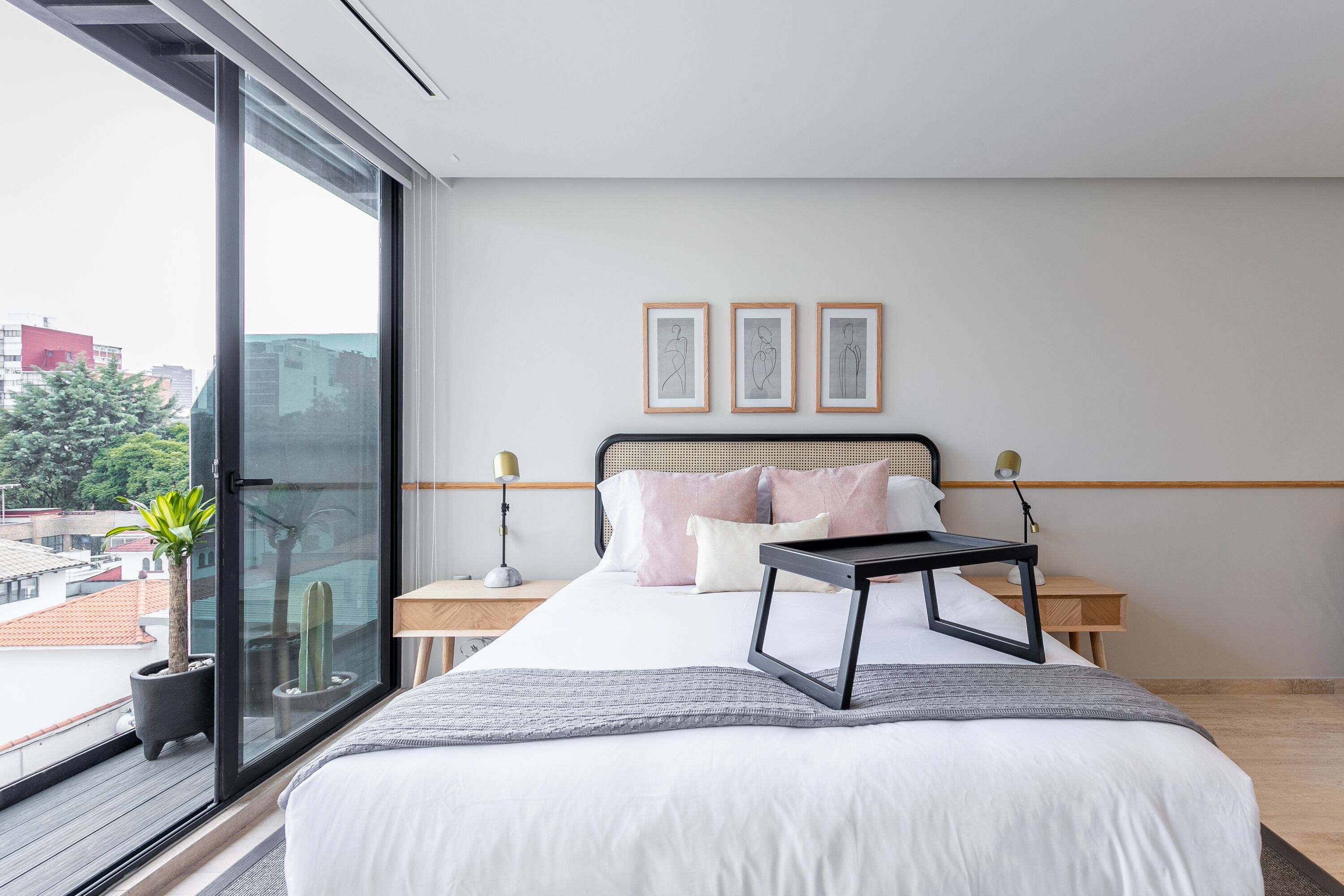 Un dormitorio moderno con un ventanal grande, una cama con una manta gris, una mesita de noche con una lámpara y una planta en maceta.