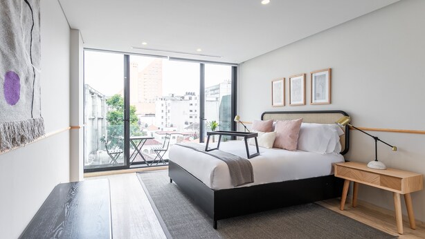 Un dormitorio moderno con una cama grande, una mesita de noche, una lámpara y vistas a la ciudad a través de amplios ventanales.