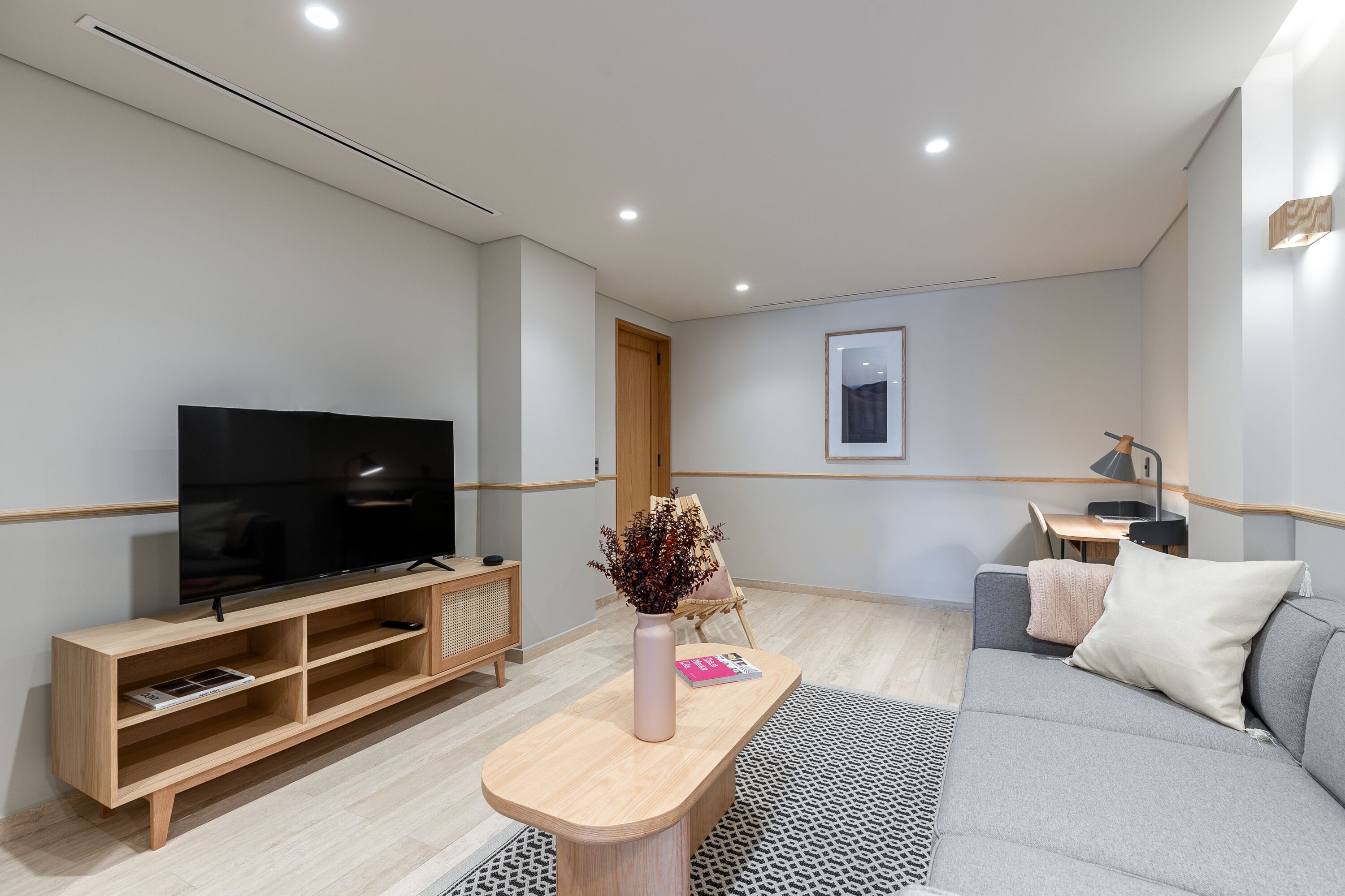 A modern living room with a flat-screen TV, wooden entertainment unit, a small table with a vase, and a grey sofa.