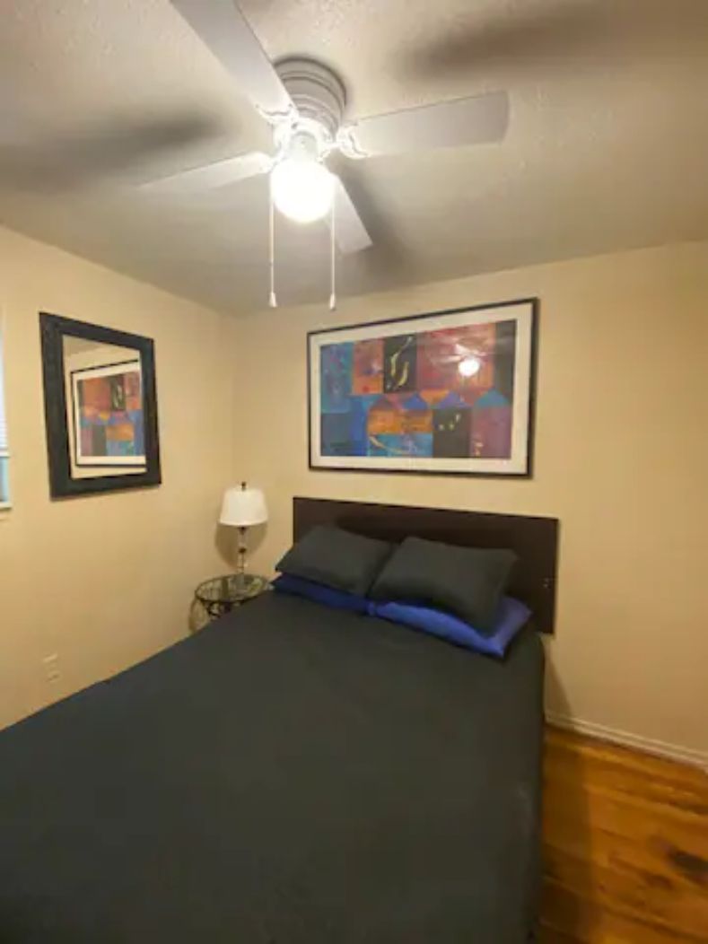 A bedroom with a bed, a ceiling fan, a nightstand, a lamp, and two framed artworks on the wall.