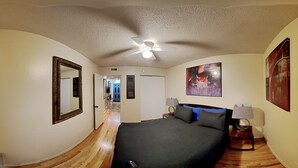 A bedroom with a bed, a ceiling fan, a painting, and a hallway leading to a door.