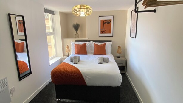 A hotel room with a bed, a bedside table, a lamp, a mirror, and artwork on the walls. - The Gateway Apartment 3 (Blackpool)