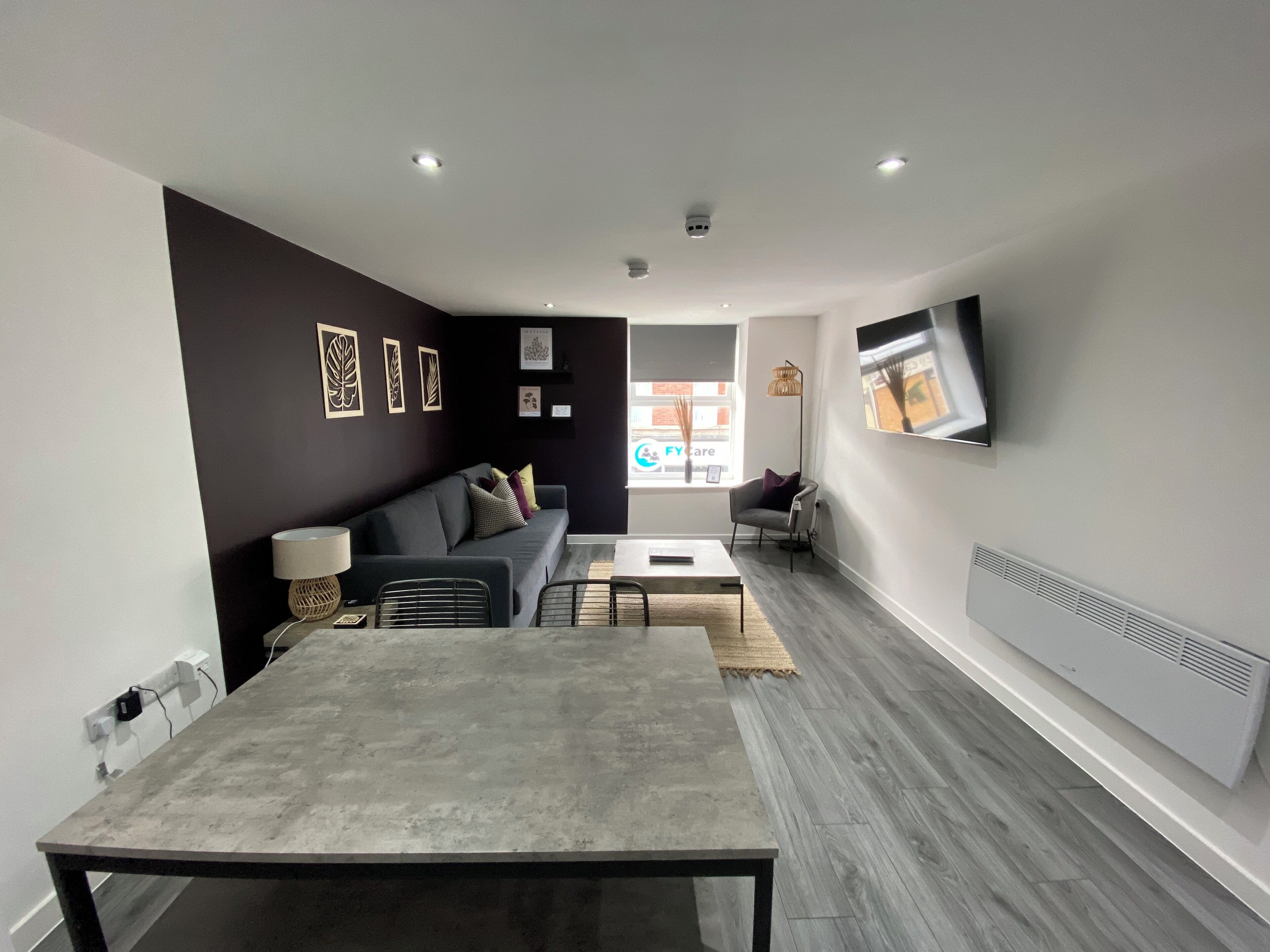 A modern living room with a grey sofa, a dining table, and a flat-screen TV mounted on the wall.