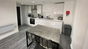 A modern kitchen with white cabinets, a built-in microwave, a stove, and a sink. There is a dining table with chairs and a fire extinguisher on the wall. - The Gateway Apartment 3 (Blackpool)