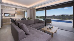 Ein modernes Wohnzimmer mit einem grauen Ecksofa, einem Holztisch und Blick auf einen Pool sowie Berge im Hintergrund.