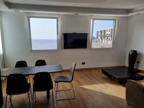 Een kamer met houten vloer, een wandgemonteerde flatscreen-tv, een eettafel met stoelen en grote ramen met uitzicht op zee en gebouwen.