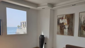 Un salon doté d’une grande fenêtre donnant sur une vue côtière, d’un canapé moderne, d’une table basse noire et de deux tableaux abstraits accrochés au mur.