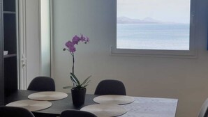 Un coin repas comprenant une table, des chaises, une plante en pot et une vue sur la mer par une fenêtre.