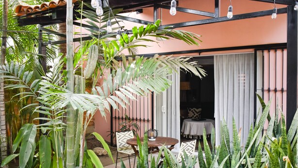 A patio area with a table and chairs, surrounded by lush greenery and a building with a tiled roof.