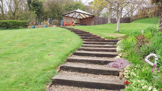 A wooden staircase leading to a wooden structure with a sloped roof, surrounded by a well-maintained garden with various plants and flowers. - The Cottage (Saltburn-by-the-Sea)