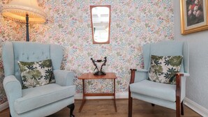 A room with floral wallpaper, two light blue armchairs with floral cushions, a small wooden table with a statue, a floor lamp, and a framed picture on the wall. - The Cottage (Saltburn-by-the-Sea)