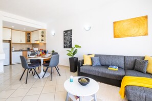 A modern living room with a grey sofa, a dining table with chairs, a small white table with a candle, and a kitchen in the background. - Cozy apartment with pool and BBQ (Albufeira)