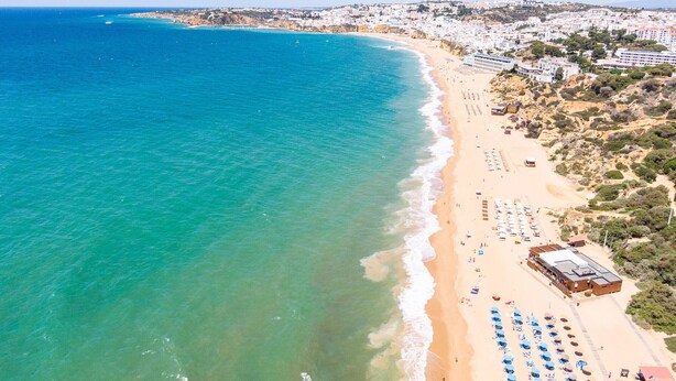 Vista aérea de un pueblo costero con playa, edificios y un mar azul cristalino.
