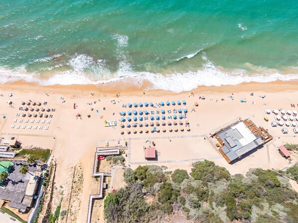An aerial view of a beach with rows of sunbeds and umbrellas, a lifeguard tower, and a building with a red roof. - Cozy apartment with pool and BBQ (Albufeira)