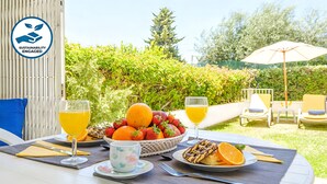 Une table dressée avec un panier de fruits, des verres de jus d’orange, une assiette de pâtisseries et une tasse, le tout installé à l’extérieur avec vue sur un jardin.