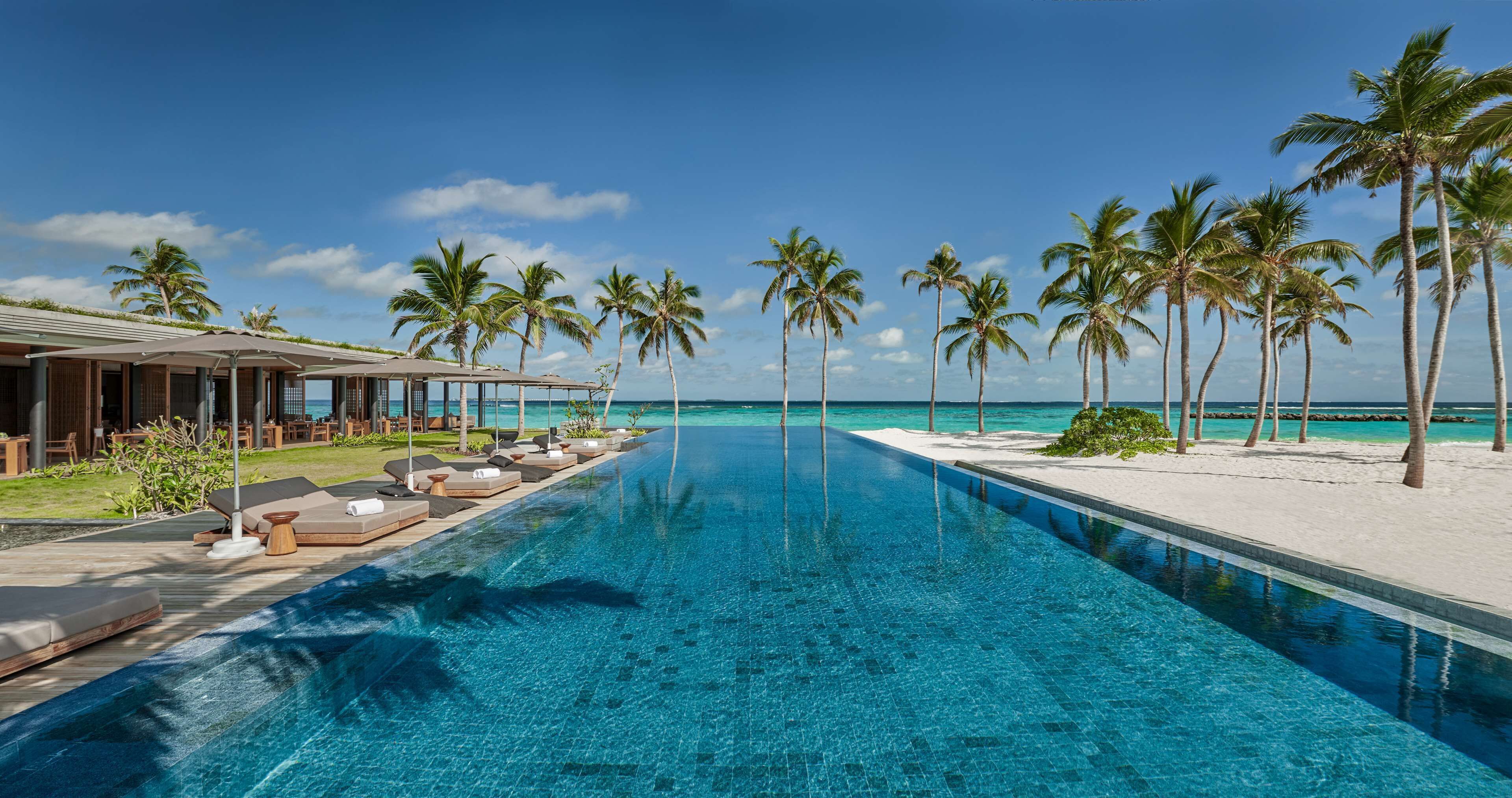 A clear blue pool with lounge chairs and umbrellas, surrounded by palm trees and a beach view.
