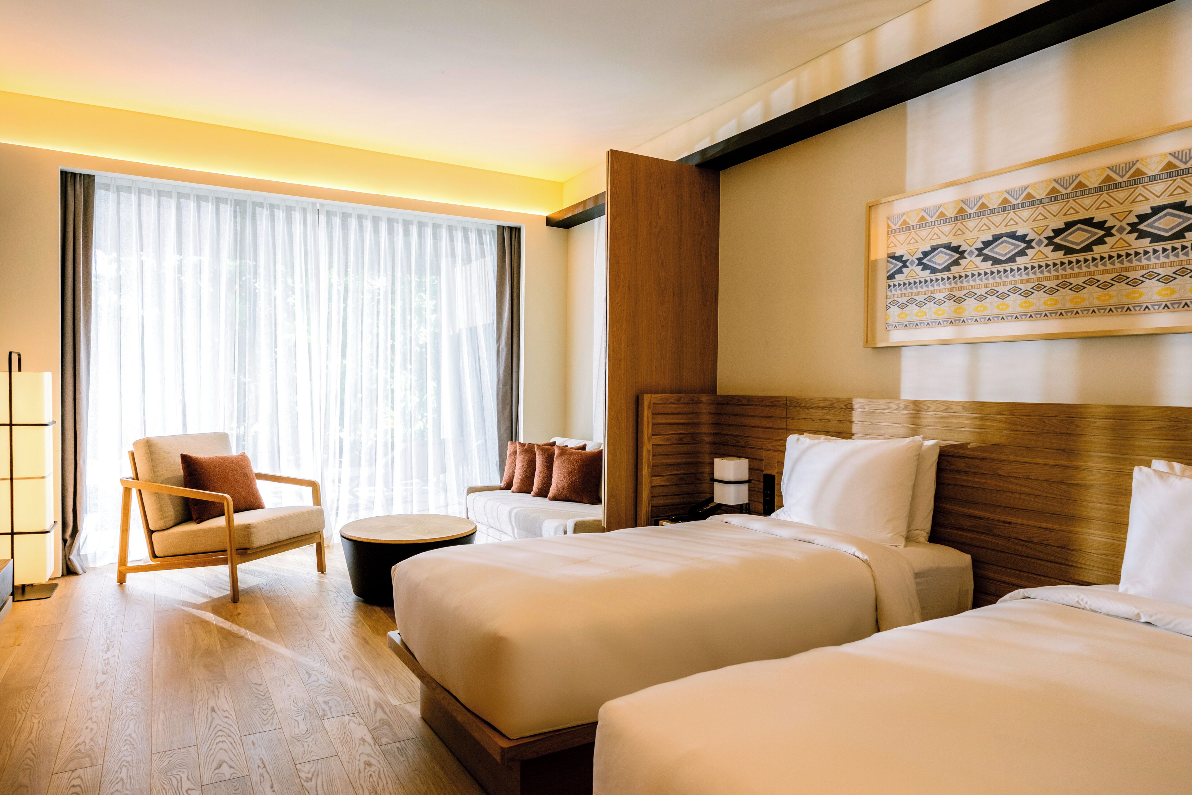 Quarto de hotel com duas camas, um espaço de estar com uma cadeira e uma mesa pequena, uma janela grande com cortinas e um objeto decorativo na parede.