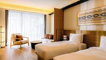 Quarto de hotel com duas camas, um espaço de estar com uma cadeira e uma mesa pequena, uma janela grande com cortinas e um objeto decorativo na parede.