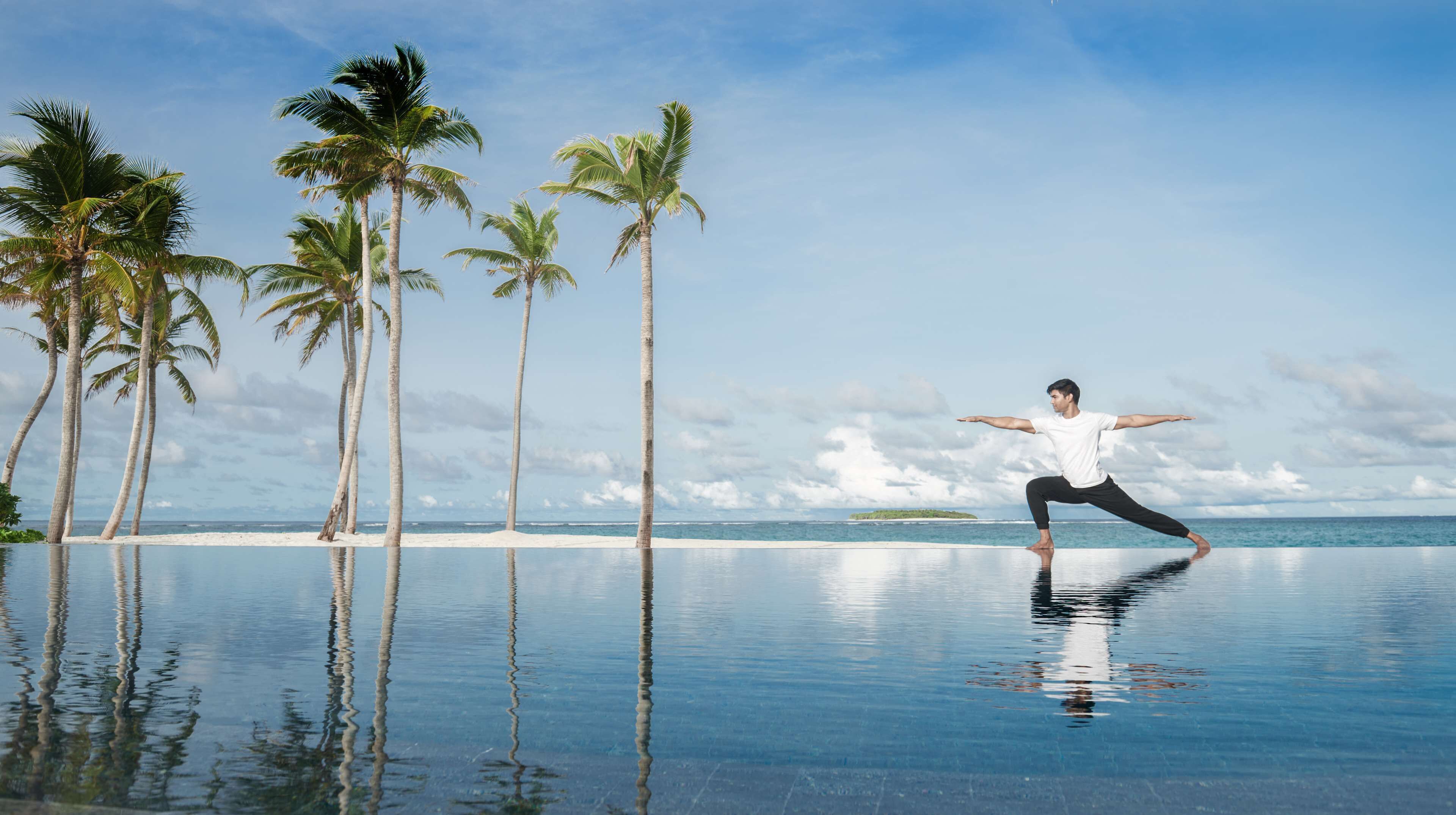 A person performing yoga on a reflective infinity pool with palm trees and a beach in the background.