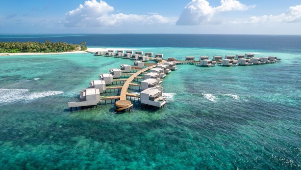 Un resort sobre el agua con varias cabañas conectadas por pasarelas, situado en aguas turquesas cristalinas y con vistas a una isla tropical y el océano.