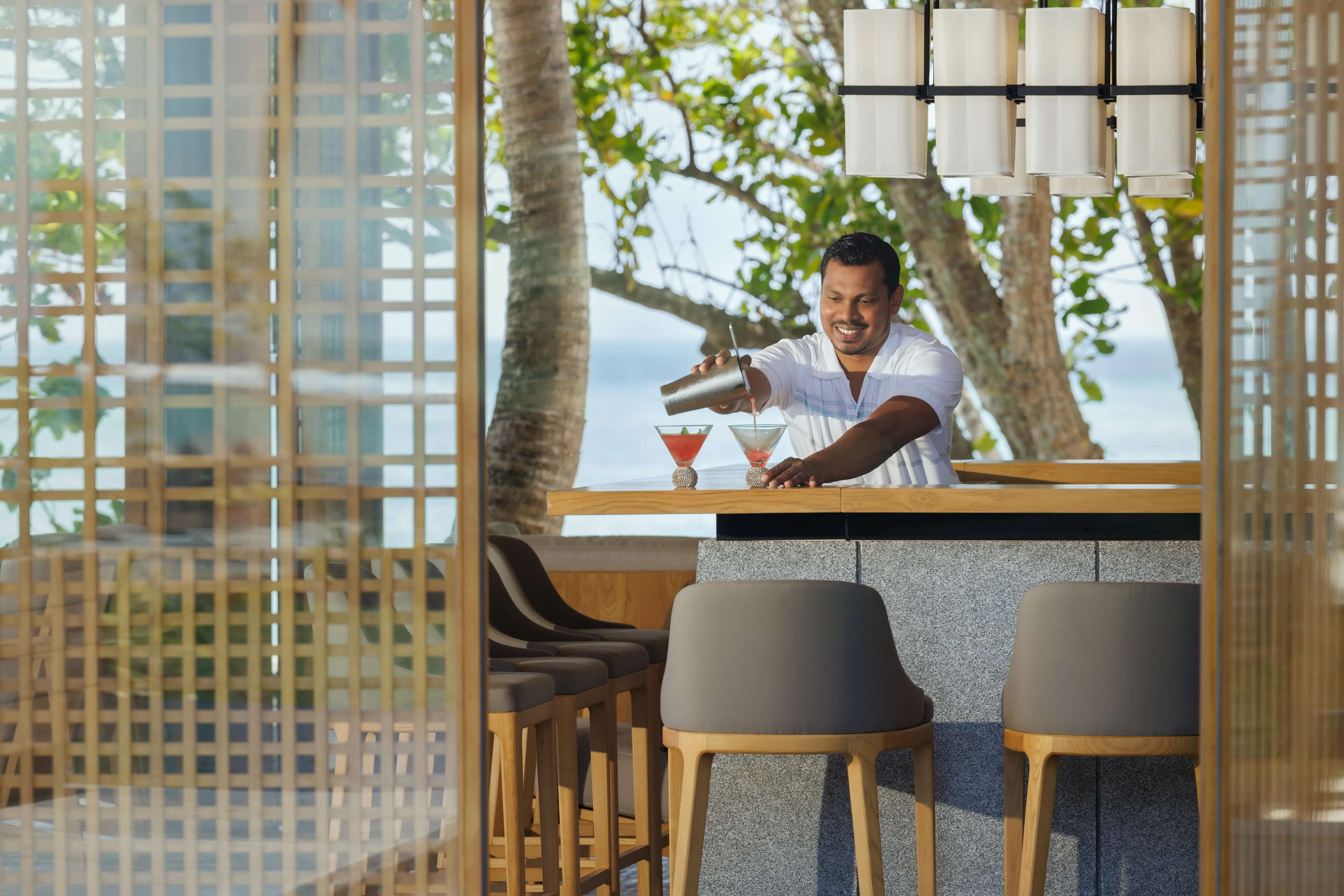 A bar area with a bartender preparing drinks, wooden chairs, and a view of the sea.
