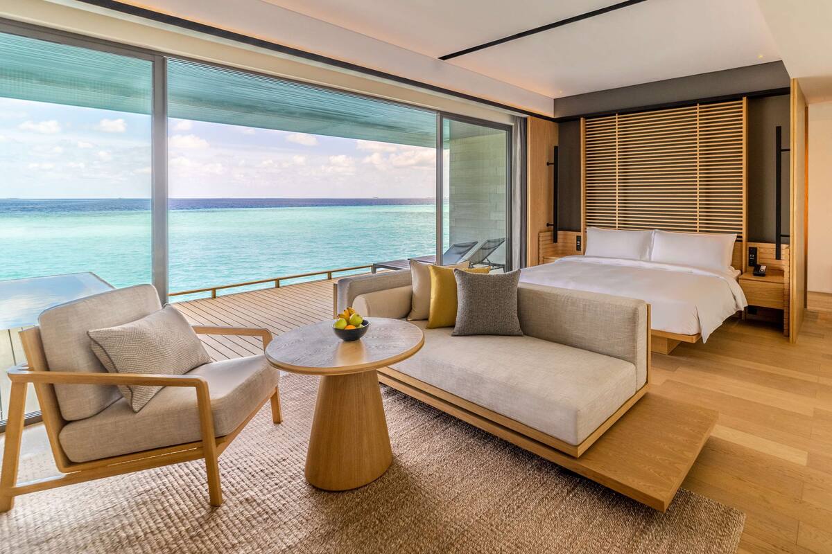 Habitación de hotel moderna con un ventanal grande que ofrece vistas al océano, una cama, un sofá, una mesita y una silla.