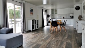 En moderne stue med en grå sofa, et spiseområde med bord og stole, et køkken med hvide skabe og hårde hvidevarer, samt et stort vindue med gardiner.