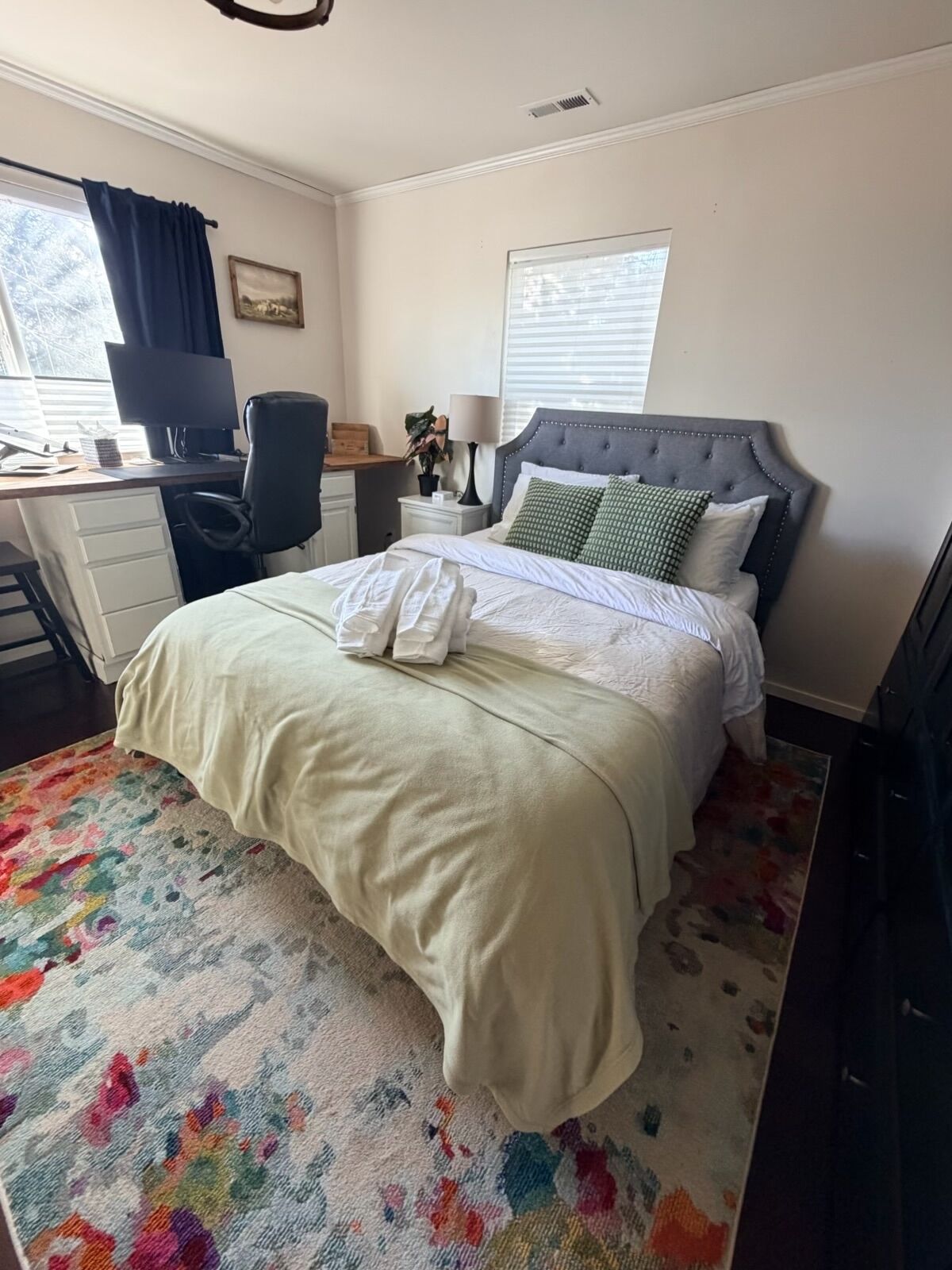 Un dormitorio con cama, escritorio con computadora, silla, mesita de noche con lámpara y ventana con cortinas.