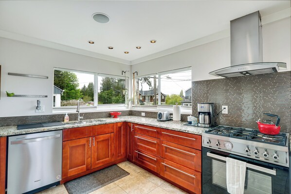 A modern kitchen with wooden cabinets, a stainless steel dishwasher, a gas stove, and a large window providing a view of the outdoors. - 4bd w/ Lake Views and Chef's Kitchen (Seattle)