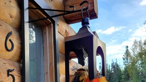 A wooden cabin with a window, a black metal number '656' on the wall, and a hanging lantern with autumn leaves and a pumpkin.