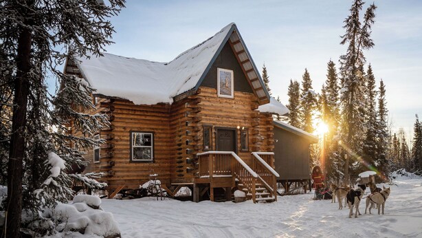 A log cabin with a snow-covered roof, wooden exterior, and a porch with steps leading to the entrance.