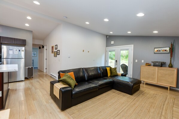 A modern living room with a black leather sectional sofa, wooden flooring, a kitchen area with a refrigerator, and a rattan cabinet. - Modern, pet friendly1/3 acre home just 25 minutes to downtown Portland (Gladstone)