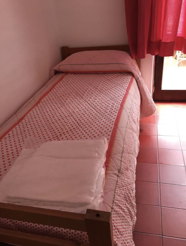 Una cama individual con colcha y almohadillas rojas y blancas a lunares, cabecera de madera y ventana con cortinas rojas.