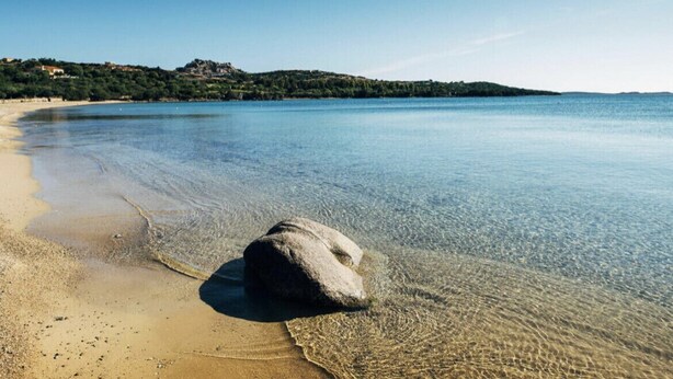 Una playa despejada con una roca grande al frente, agua tranquila y una costa distante con edificios.