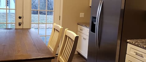 A kitchen with a wooden table, chairs, a refrigerator, and a window with a view of the outdoors.
