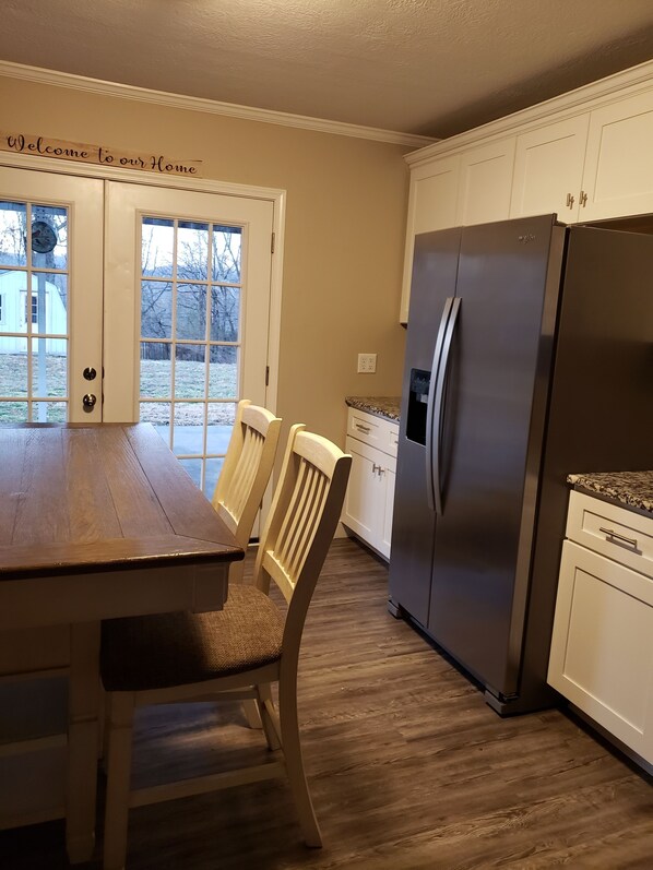 A kitchen with a wooden table, chairs, a refrigerator, and a window with a view of the outdoors.
