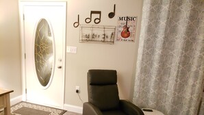 Una habitación con un sillón verde oscuro, una mesita redonda pequeña, una cortina estampada y una pared con un cartel de temática musical.
