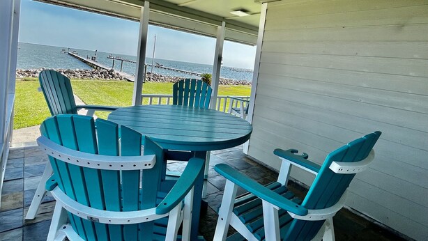 Een overdekt terras met een ronde tafel en vier stoelen, met uitzicht op een rotsachtige kustlijn en een pier.