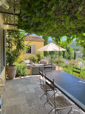 Una veranda coperta all'aperto con tavolo e sedie, immersa nel verde con una casa sullo sfondo.