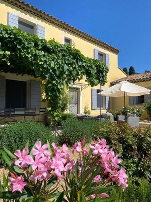 Un edificio giallo con tetto in tegole, un portico coperto con mobili da esterno e un giardino con fiori rosa.