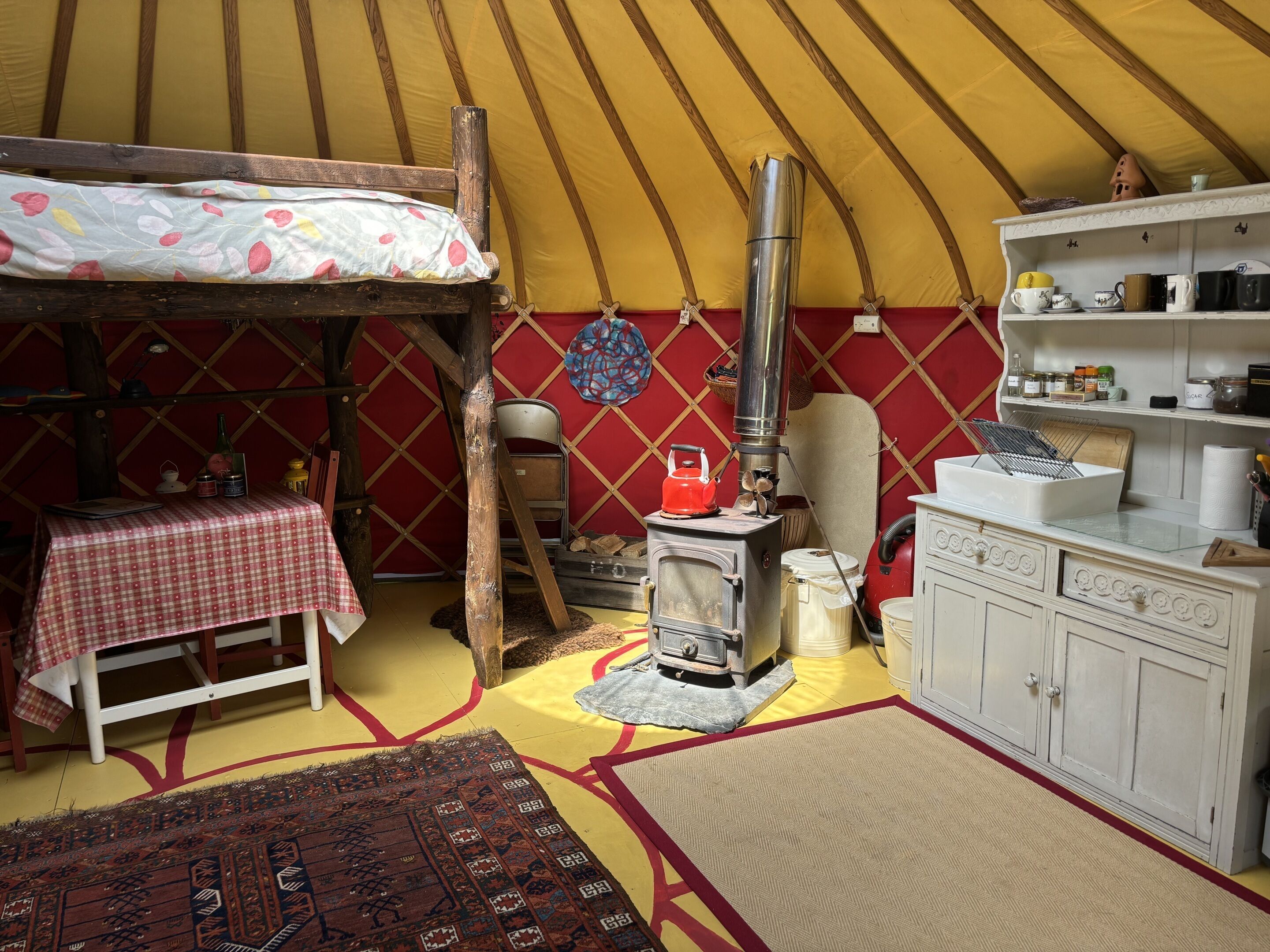 A cozy interior with a bed, a table, a stove, and a shelf with kitchen items.